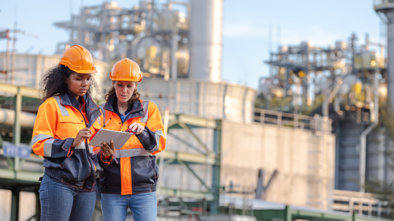 Two women in high-visibility safety jackets and orange hard hats review information on a tablet at an industrial facility, highlighting workforce roles in water or utility infrastructure.