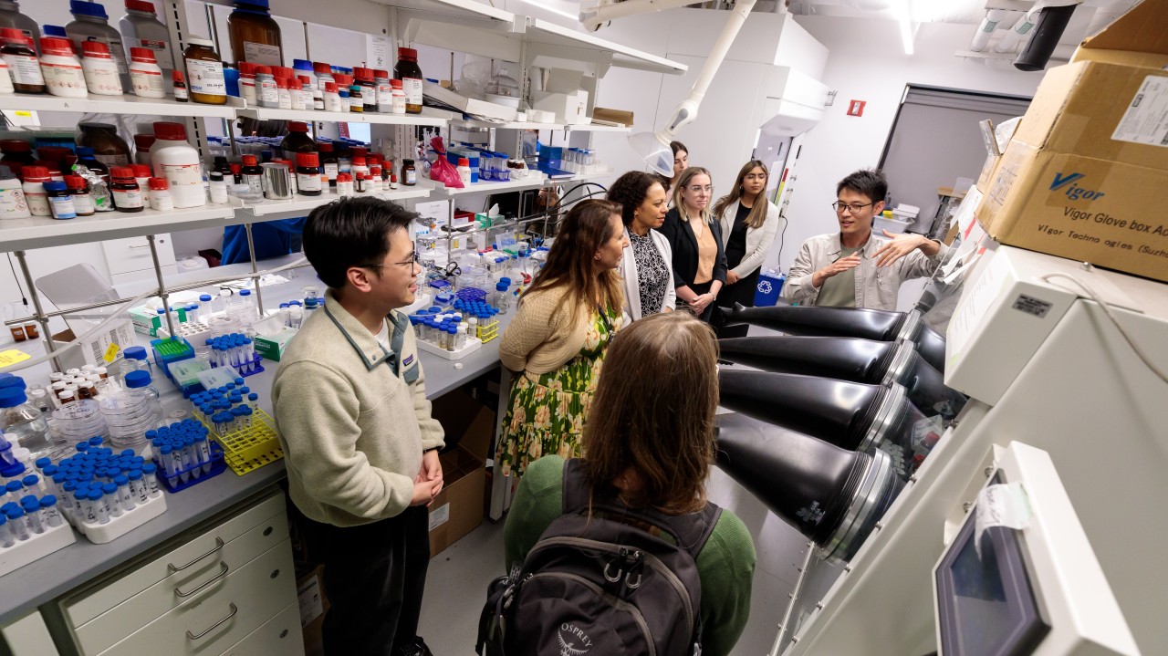A group of people gathers in a laboratory as a researcher explains equipment and experiments related to chemical processes. Shelves filled with test tubes, beakers, and chemical containers line the workspace.